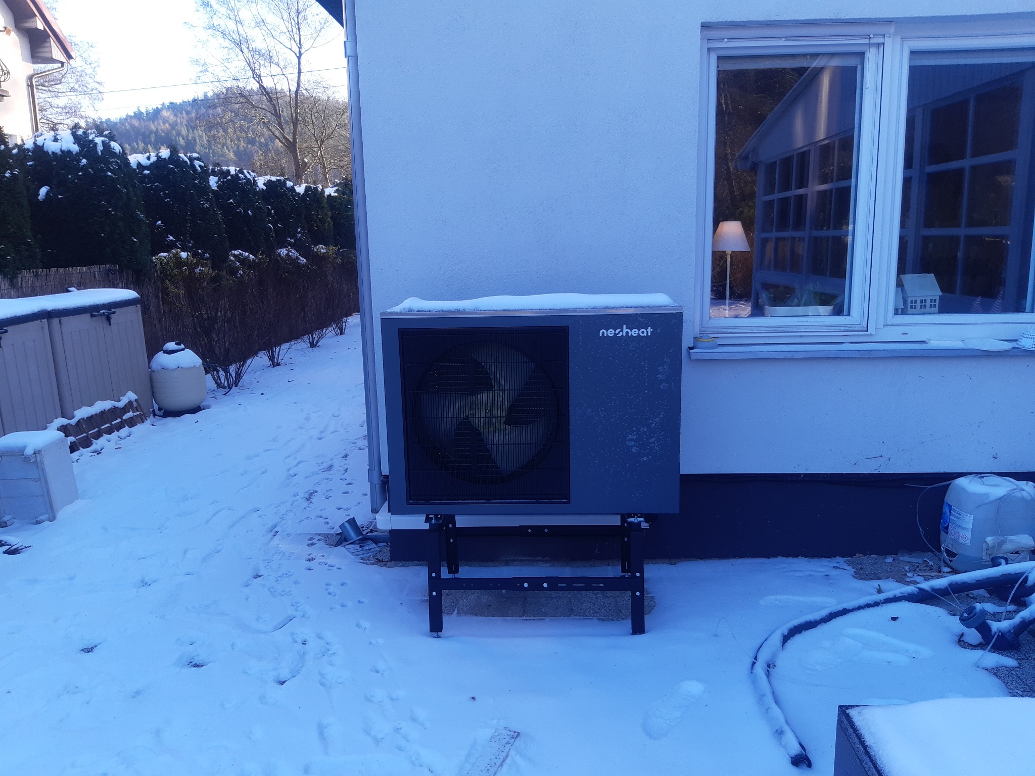 Szara pompa ciepła marki Nesheat zamontowana na czarnej metalowej konstrukcji, stoi na śniegu przy białej ścianie budynku z oknem, widoczne ślady na śniegu i rury instalacyjne.