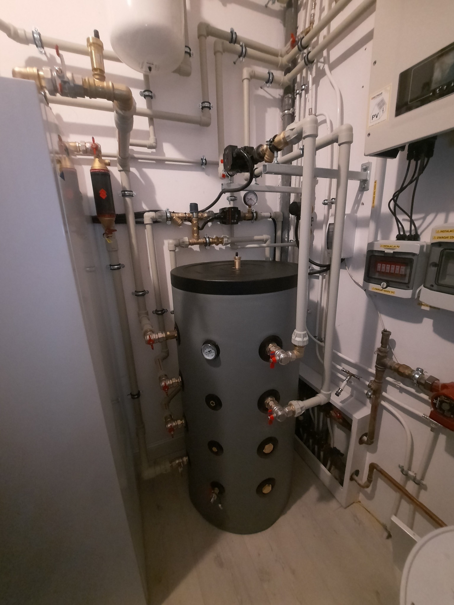 Szary zbiornik buforowy ciepła z licznymi rurami i zaworami w kotłowni, widoczny panel fotowoltaiczny i manometry.