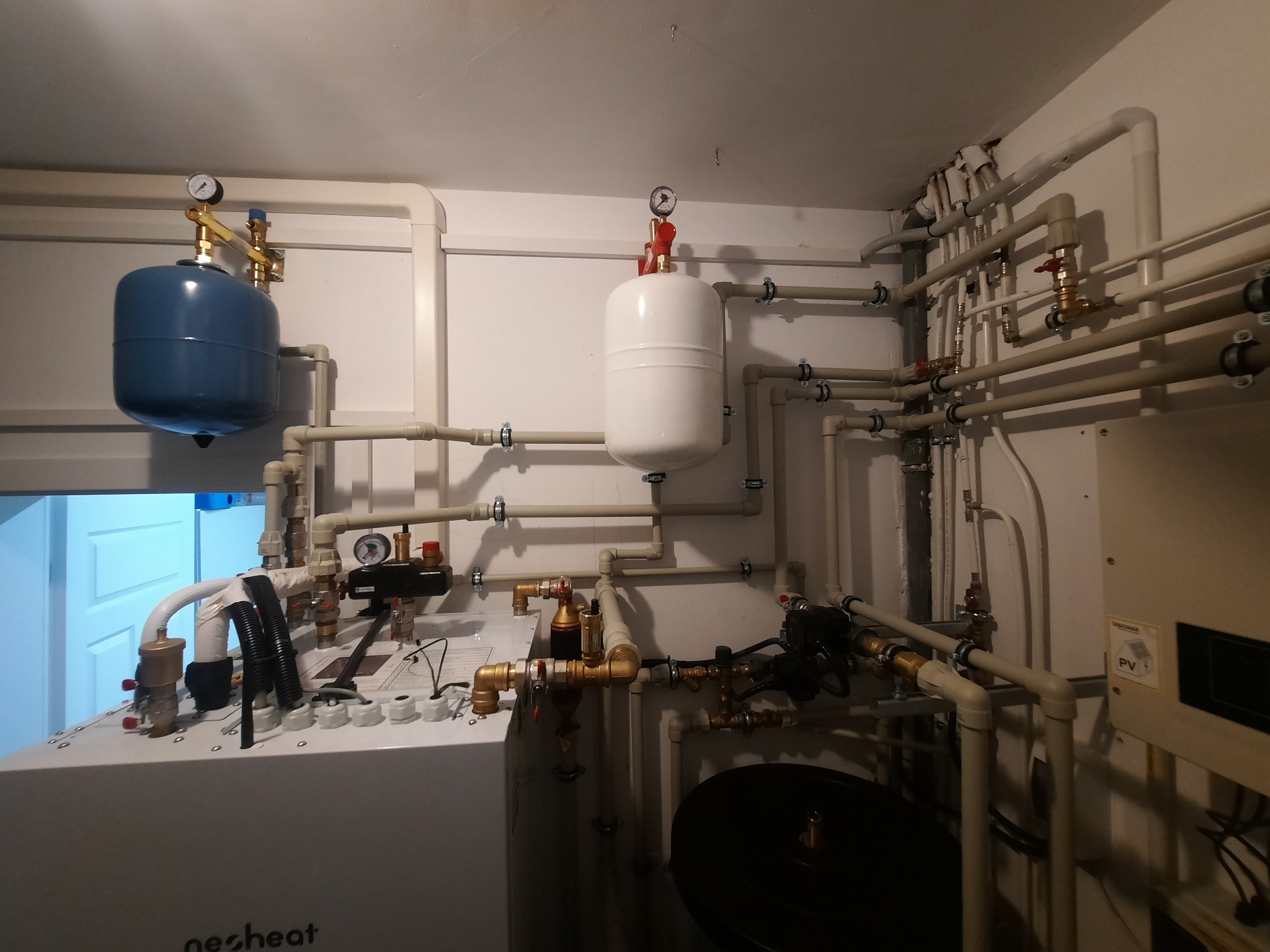 Kompleksowa instalacja grzewcza w kotłowni z zbiornikami wyrównawczymi, rurami i zaworami, widoczne manometry i elementy sterujące, białe ściany w tle.