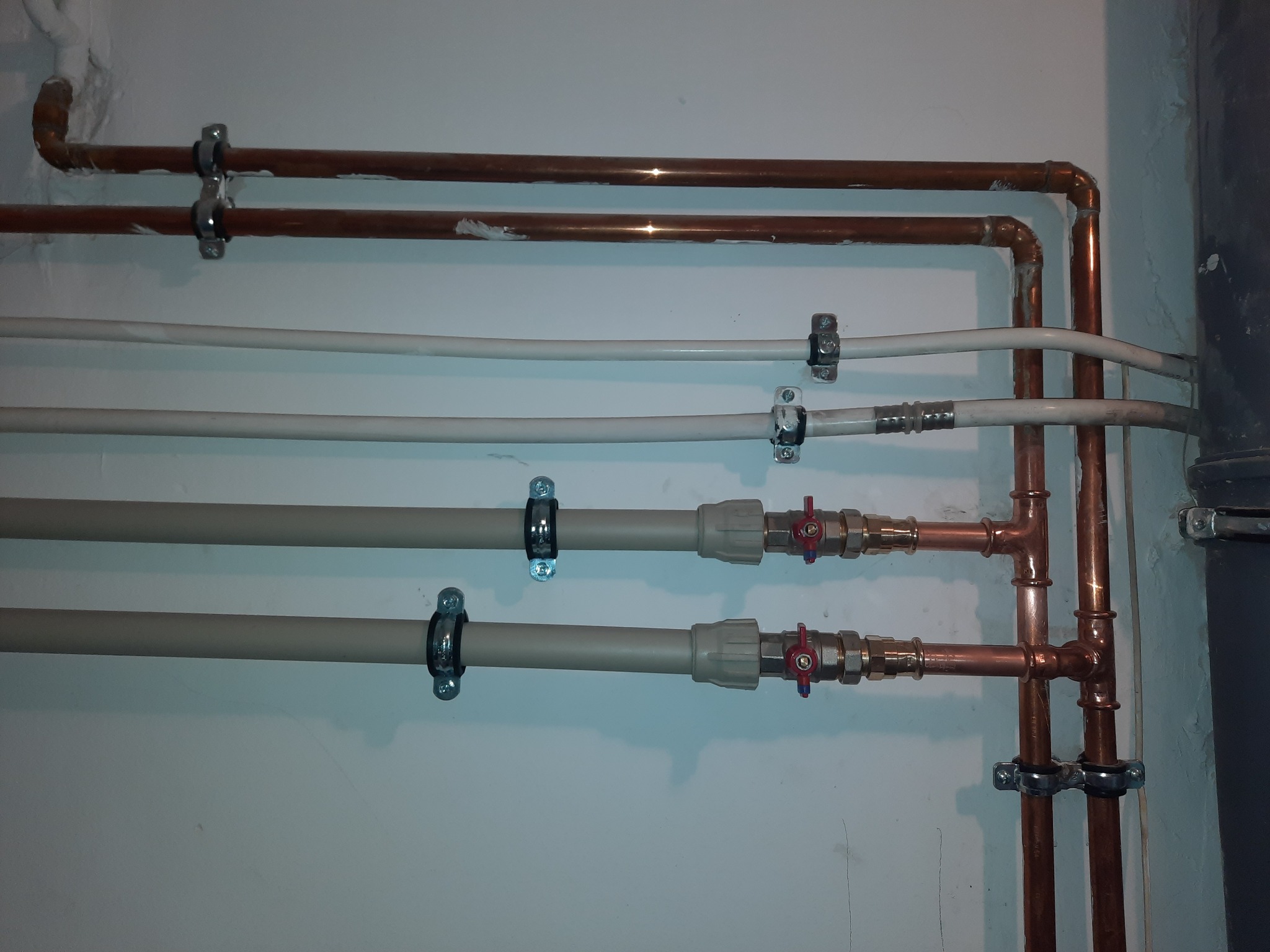 Instalacja rur miedzianych i plastikowych z zaworami, zamocowanych do ściany za pomocą metalowych obejm.