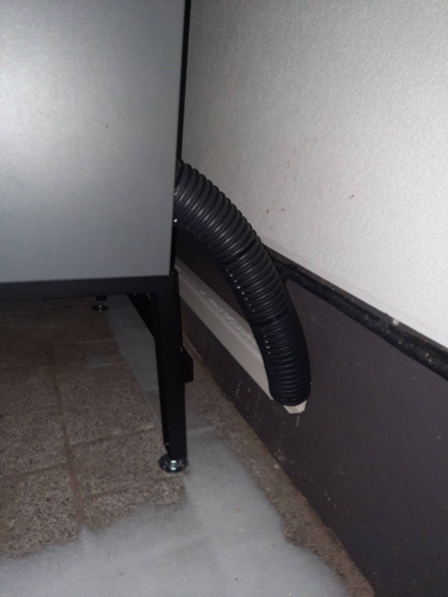 Szara obudowa urządzenia z czarnym karbowanym przewodem odprowadzającym, ułożonym wzdłuż ściany z szarą lamperią, widoczne płytki podłogowe.