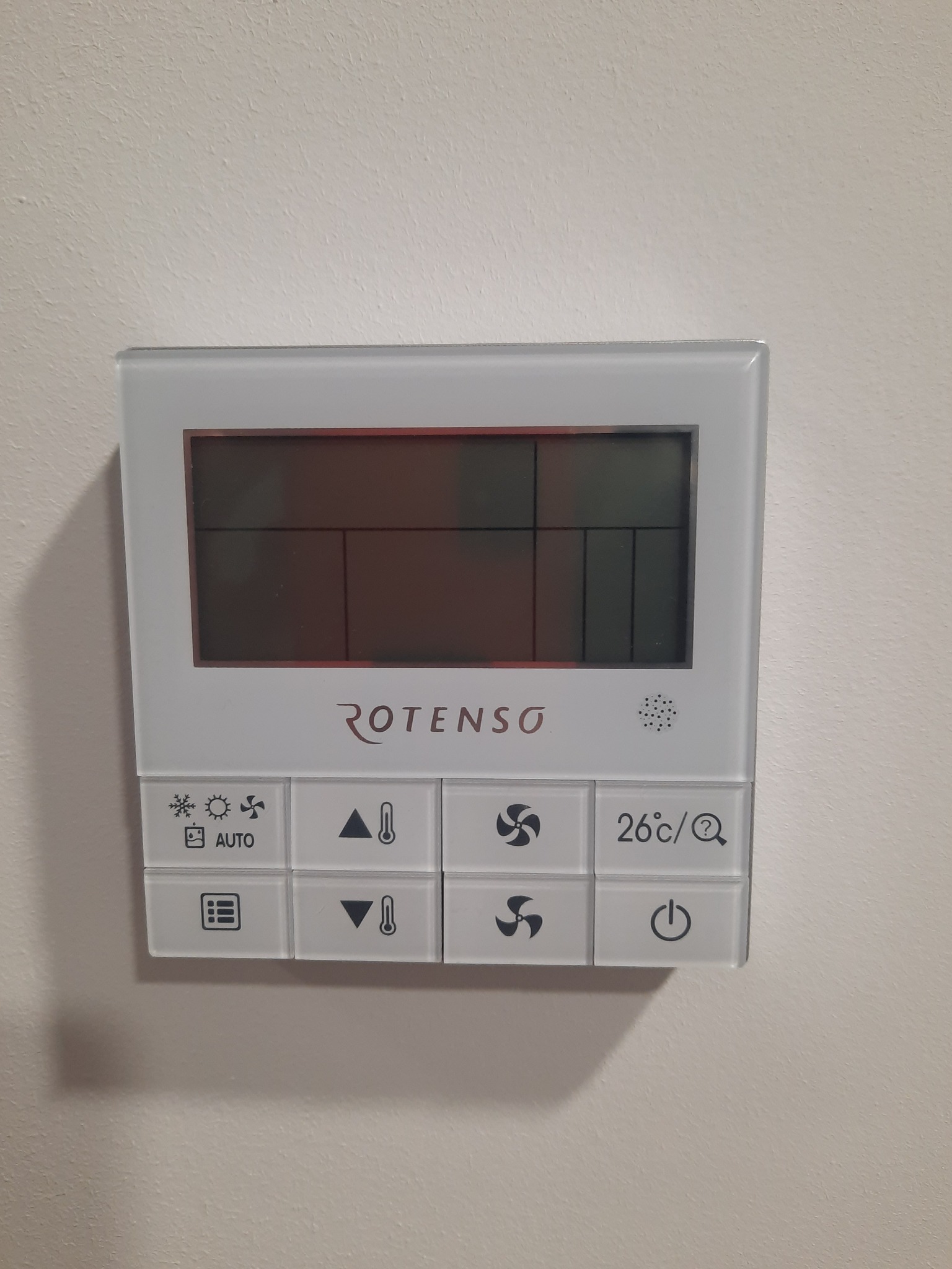 Biały, szklany panel sterowania klimatyzacją marki Rotenso, zamontowany na kremowej ścianie. Wyświetlacz jest wyłączony, a przyciski pokazują różne funkcje, takie jak temperatura, wentylator i tryb...