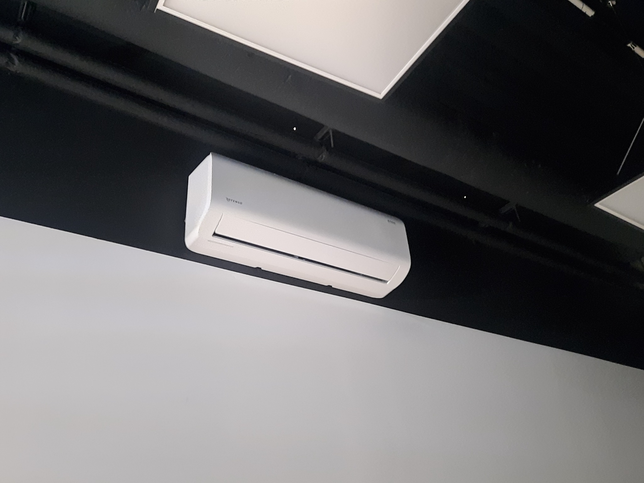Biała jednostka klimatyzacji zamontowana pod czarnym sufitem z widocznymi instalacjami, na tle białej ściany.