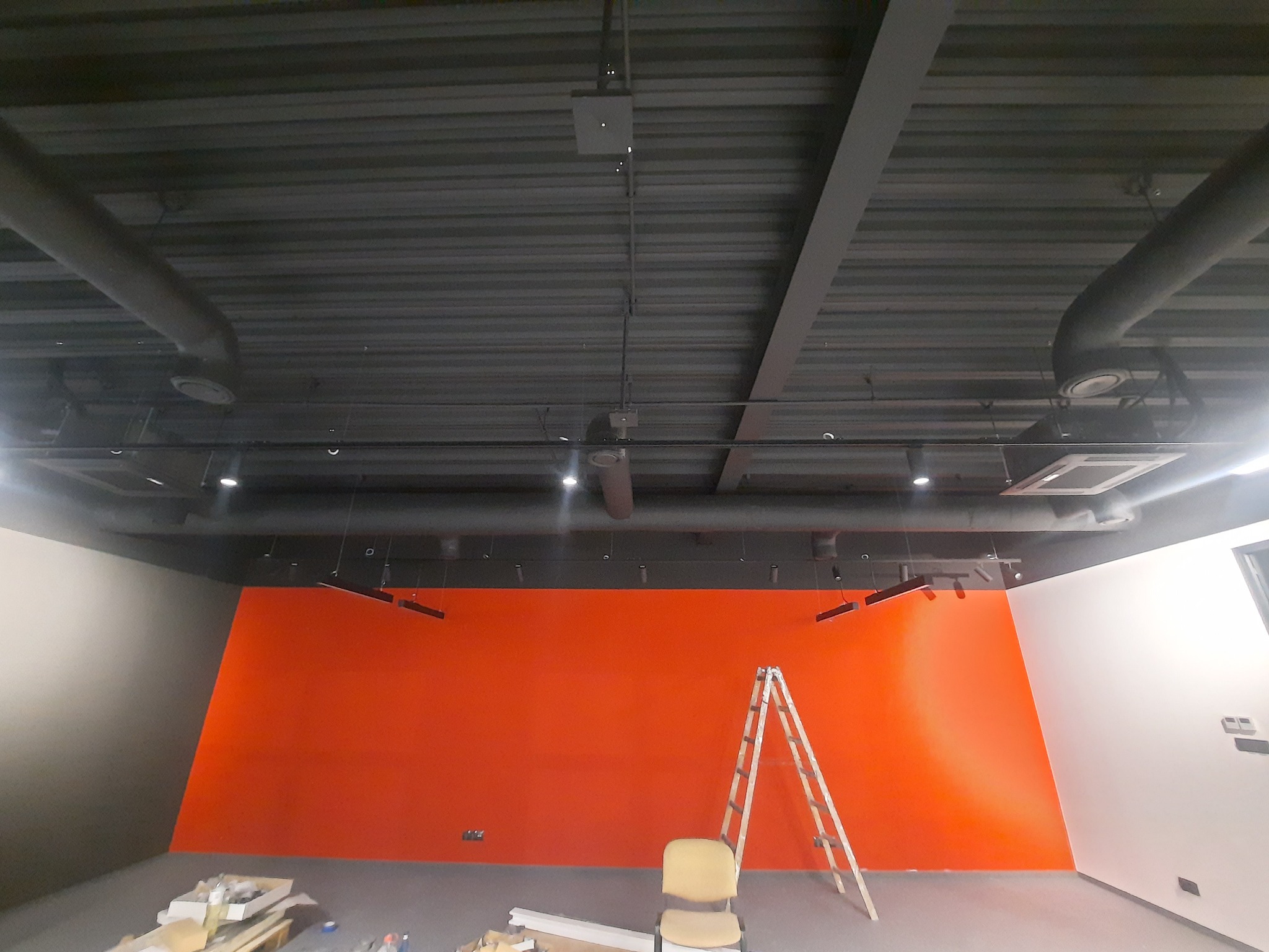 Wnętrze biurowe w trakcie wykańczania, z widoczną pomarańczową ścianą, drabina, krzesło, elementy instalacji pod sufitem oraz materiały budowlane na podłodze.