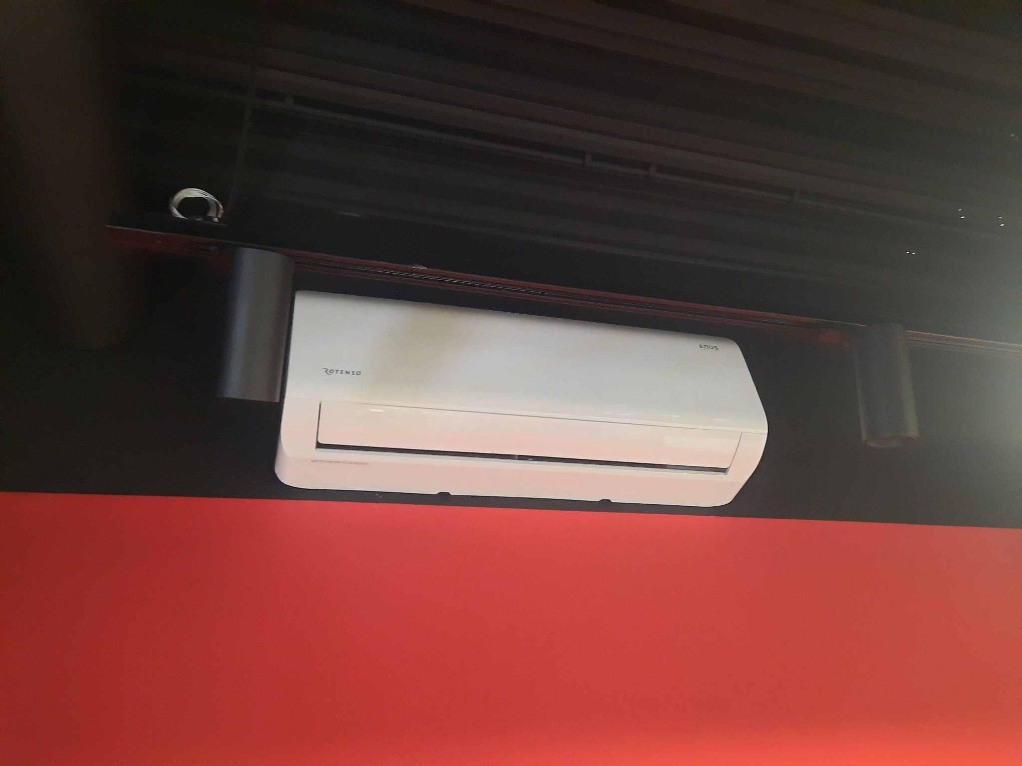 Biały klimatyzator marki Rotenso zamontowany pod sufitem z widoczną kamerą monitoringu, na tle czerwonej ściany.