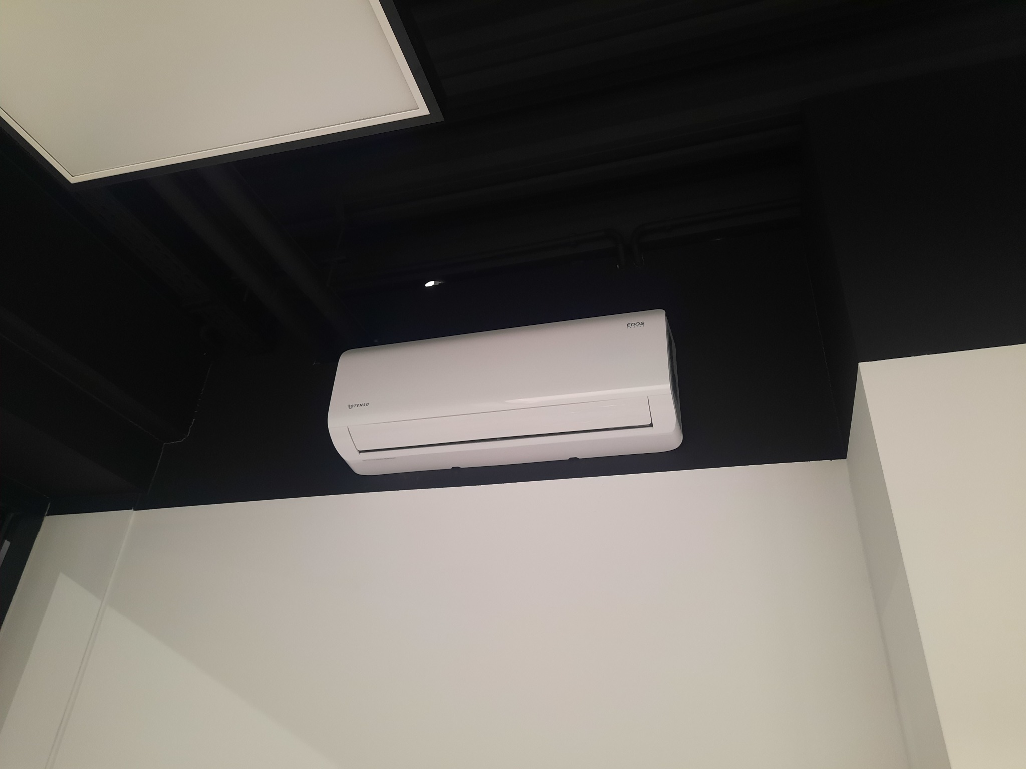 Biały klimatyzator marki Rotenso zamontowany na ścianie w pomieszczeniu z czarnym sufitem i białą ścianą, widok z dołu.