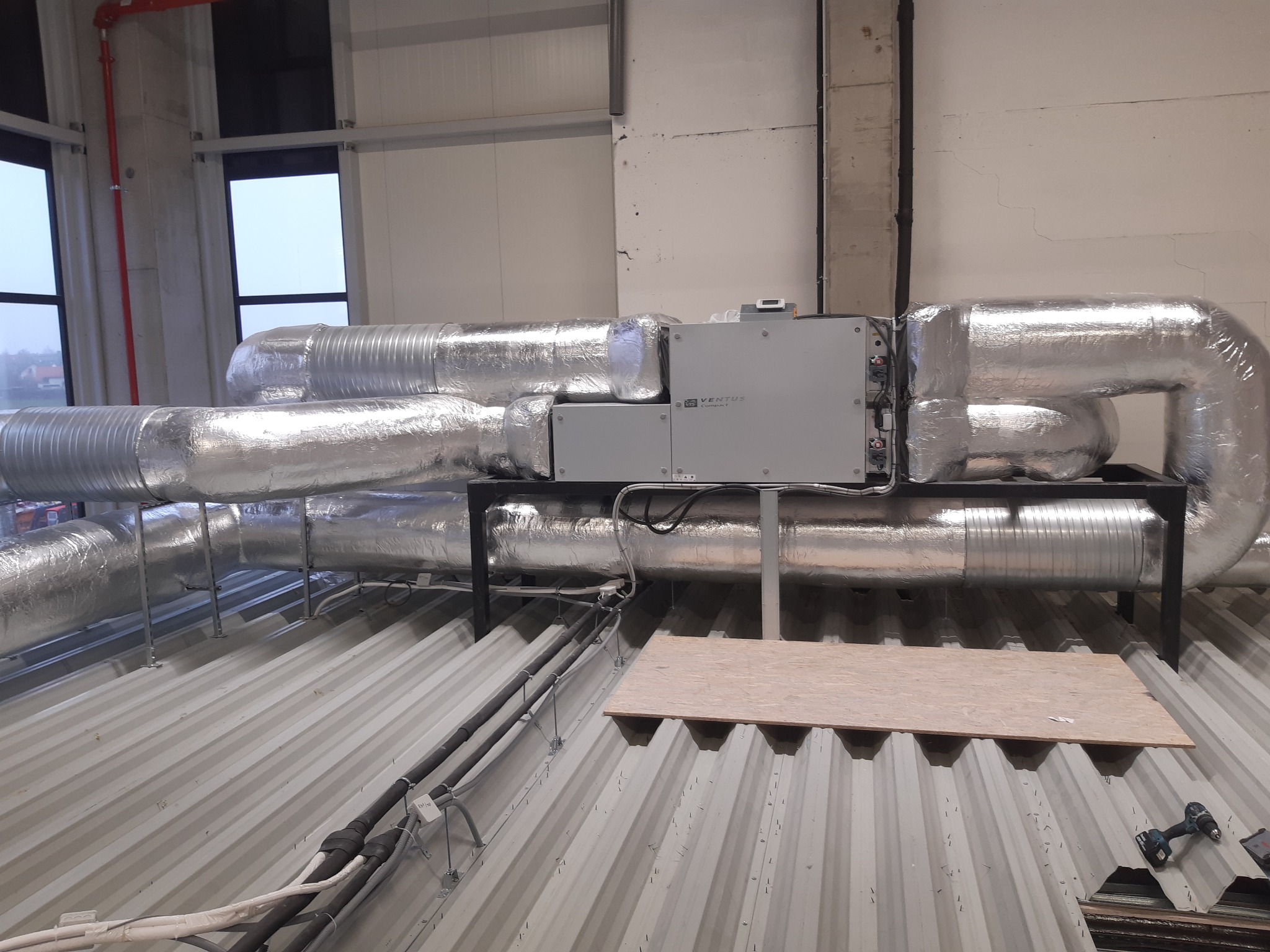 Srebrne, izolowane przewody wentylacyjne w przemysłowym budynku, podwieszone na metalowej konstrukcji, z widocznym modułem sterującym Ventus Kompakt i fragmentem płyty OSB na falistej blasze dachowej.