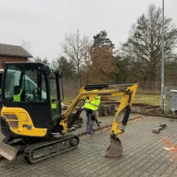 Żółta mini koparka Yanmar SV18 na kostce brukowej, pracownik w żółtej kamizelce używa wiertarki udarowej do przygotowania wykopu pod kable, obok ułożone kostki brukowe.