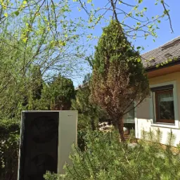 Zewnętrzna jednostka pompy ciepła Hewalex z dwoma wentylatorami, zainstalowana przy budynku jednorodzinnym, otoczona zielenią i iglakami.