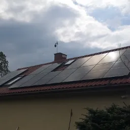 Panele słoneczne na dachu domu jednorodzinnego z czerwonej dachówki, widoczne okna dachowe i komin z kamerą monitoringu, pochmurne niebo w tle.