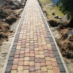 Nowo ułożony chodnik z kostki brukowej w odcieniach brązu i beżu, obrzeża z ciemnoszarej kostki, widoczne przygotowane podłoże z piasku i ziemi po bokach, brama w tle.