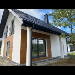 Nowoczesny dom jednorodzinny z białą elewacją i elementami dekoracyjnymi z drewnopodobnych paneli, czarny dach z blachy na rąbek stojący, widoczne orynnowanie i komin.