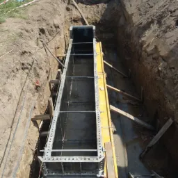 Metalowa szalunek do fundamentów w wykopie, zabezpieczona drewnianymi podporami, widoczne ślady wody i błota.