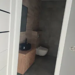 Nowoczesna łazienka z umywalką nablatową w kolorze czarnym i szafką z drewnianymi lamelami, widok przez białe drzwi z czarnym akcentem, szare płytki na ścianach.