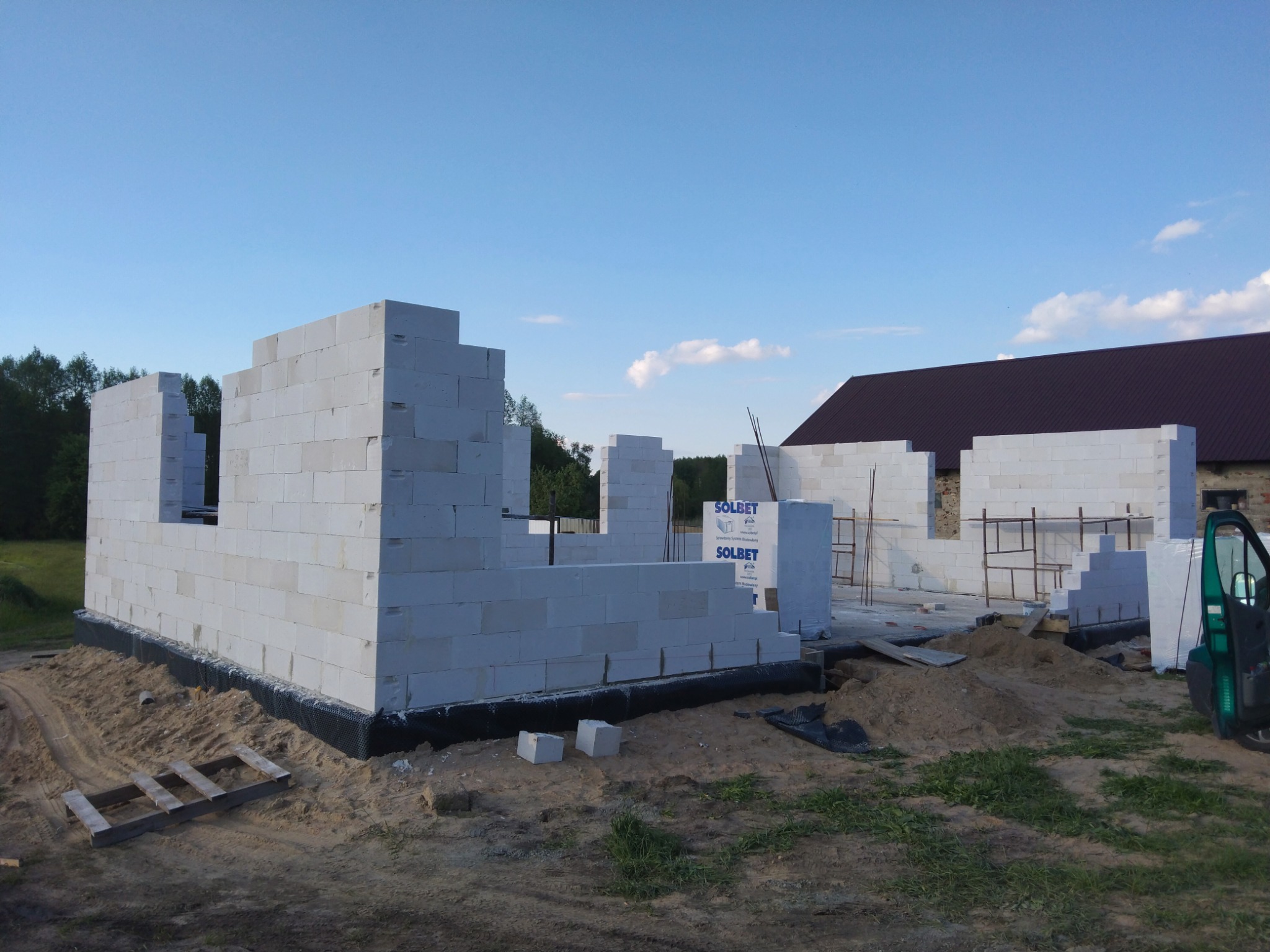 Wznoszenie ścian parteru domu z białych bloczków na fundamencie z izolacją, widoczne palety z bloczkami, teren budowy z piaskiem i zielenią, w tle budynek z brązowym dachem i las.