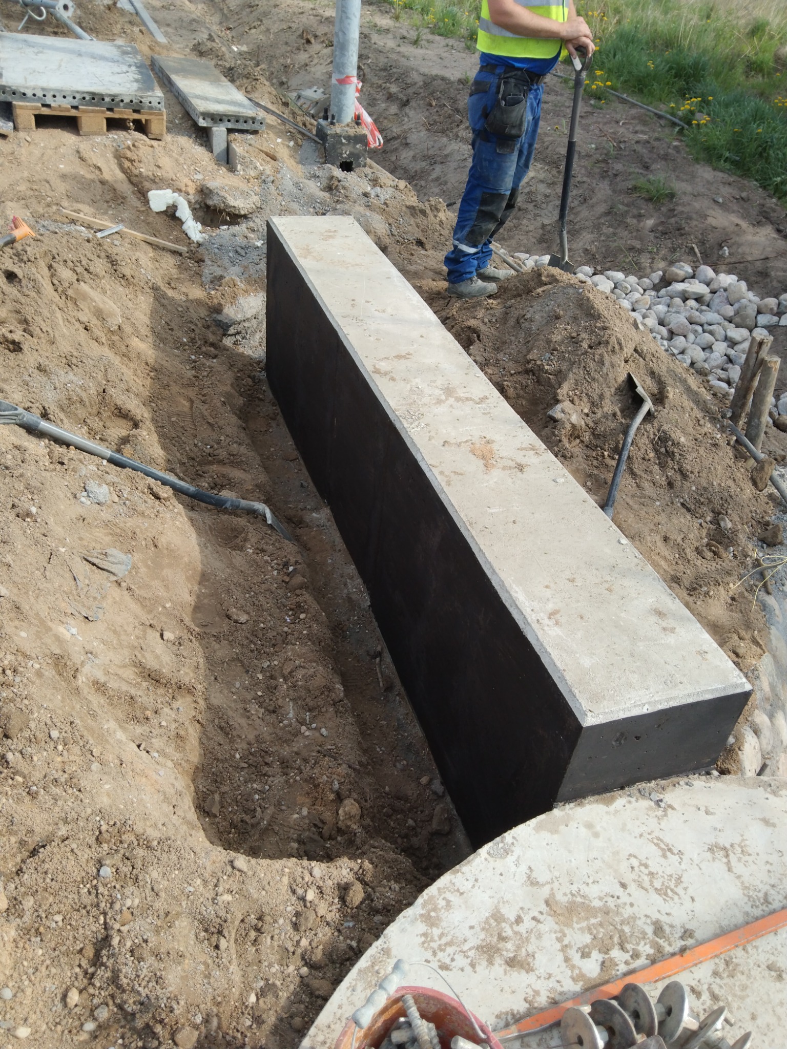 Betonowy element fundamentu, zabezpieczony czarną izolacją, umieszczony w wykopie, widoczny pracownik z łopatą w tle.