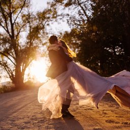 Fotograf Anna Shaulskaya - Para w objęciach, panna młoda w zwiewnej sukni tanecznej, zachód słońca w tle drzew, na piaszczystej drodze.
