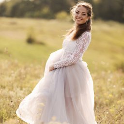 Fotograf Anna Shaulskaya - Uśmiechnięta kobieta w koronkowej sukni i zwiewnej spódnicy pozuje na łące, w tle rozmyte drzewa.