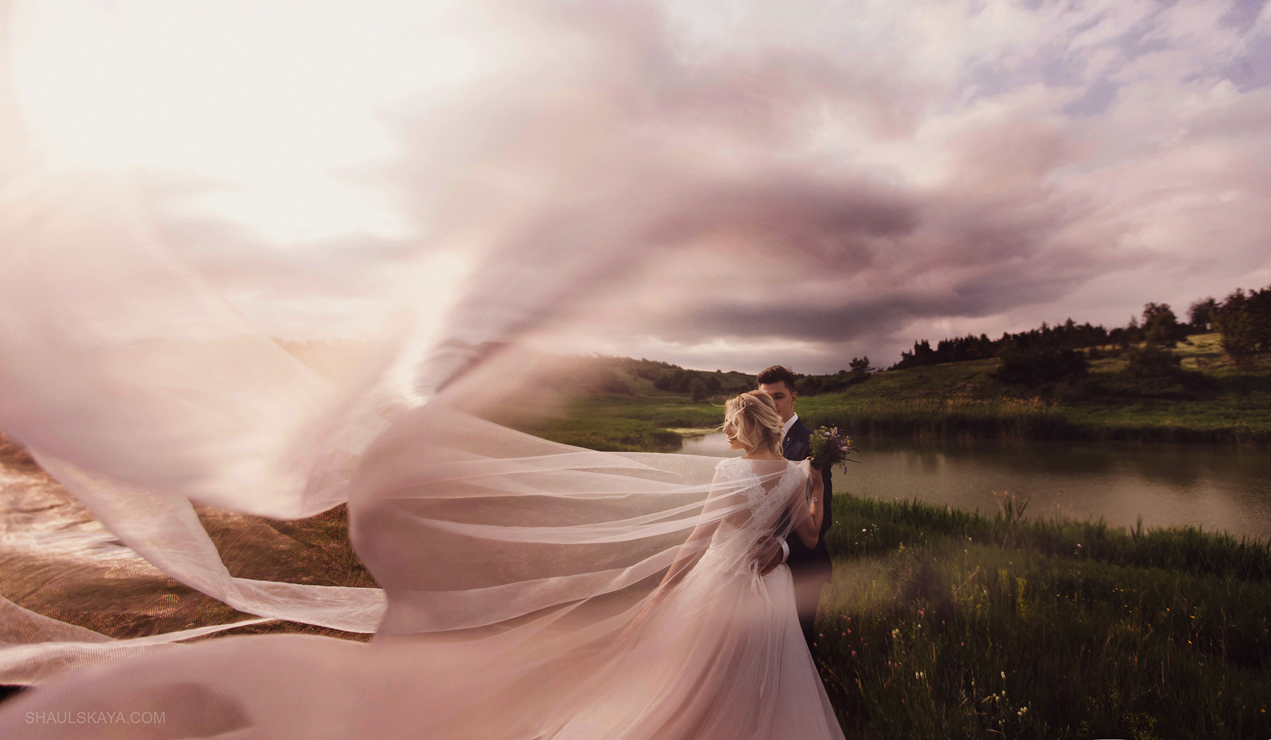 Para narzeczeńska w plenerze: panna młoda w zwiewnej sukni z długim welonem, który powiewa na wietrze, obejmowana przez pana młodego na tle łąki i jeziora.