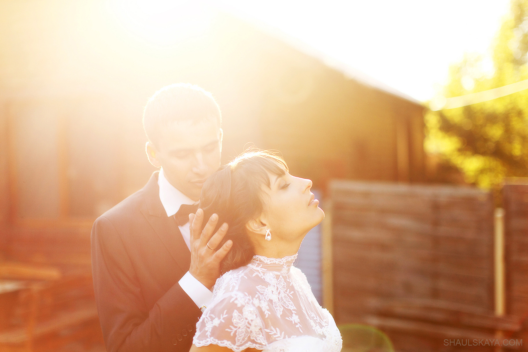 Para w ślubnych strojach w ciepłym świetle zachodzącego słońca; mężczyzna w smokingu delikatnie dotyka włosów kobiety w koronkowej sukni, która patrzy w górę.