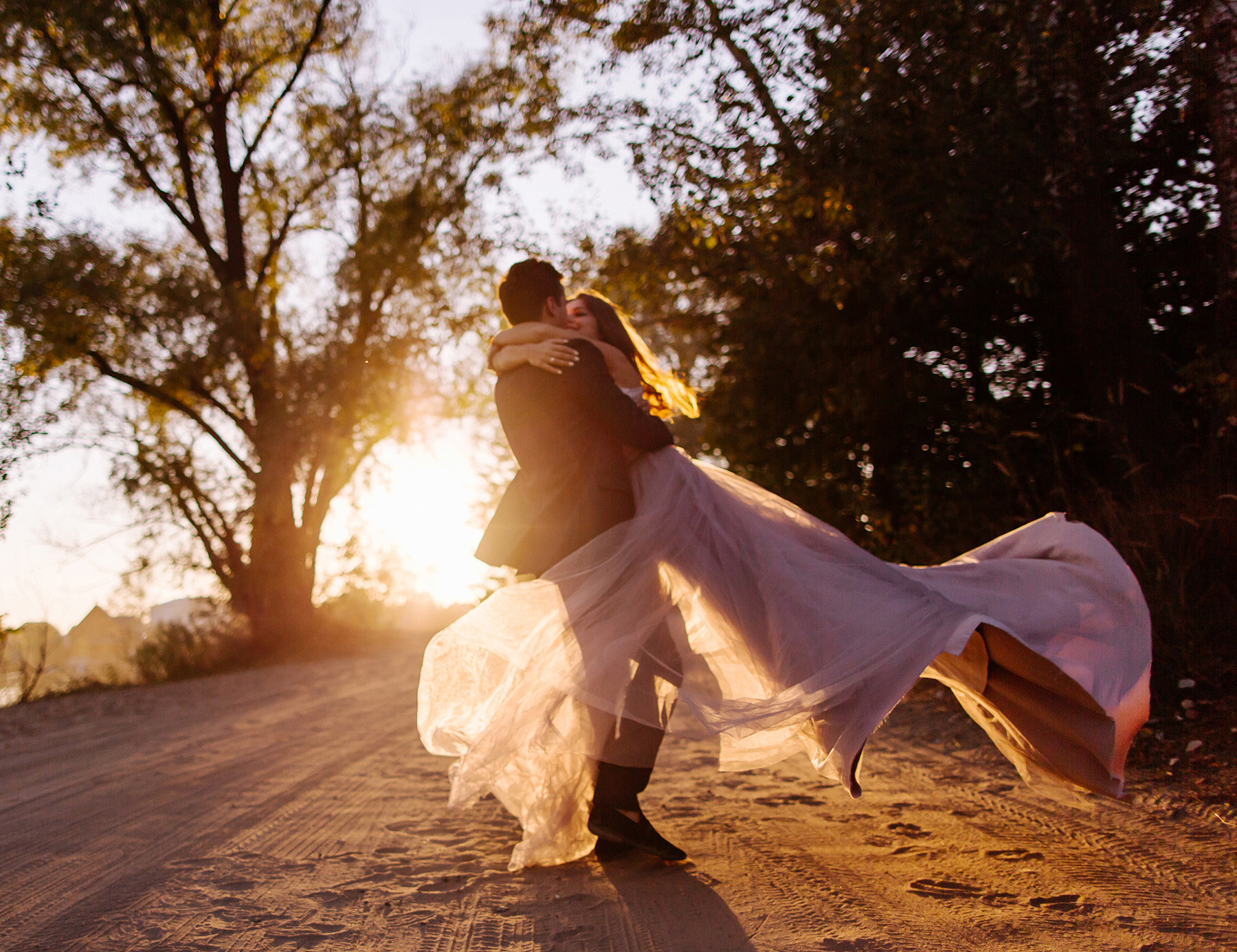 Para w objęciach, panna młoda w zwiewnej sukni tanecznej, zachód słońca w tle drzew, na piaszczystej drodze.