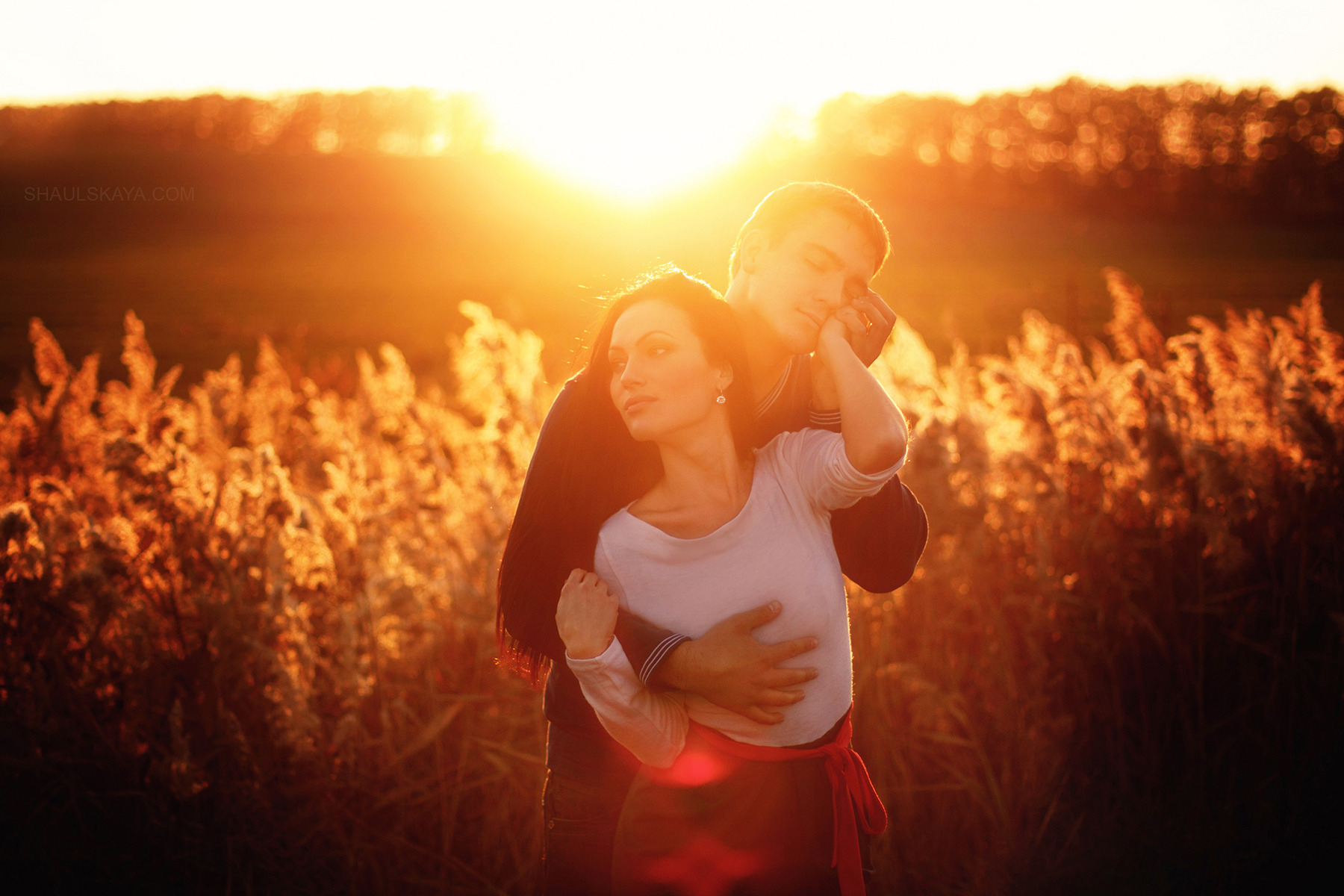 Zakochana para w objęciach na tle zachodzącego słońca i wysokich traw, kobieta w białej bluzce i spódnicy z czerwoną szarfą, mężczyzna w ciemnej bluzie.