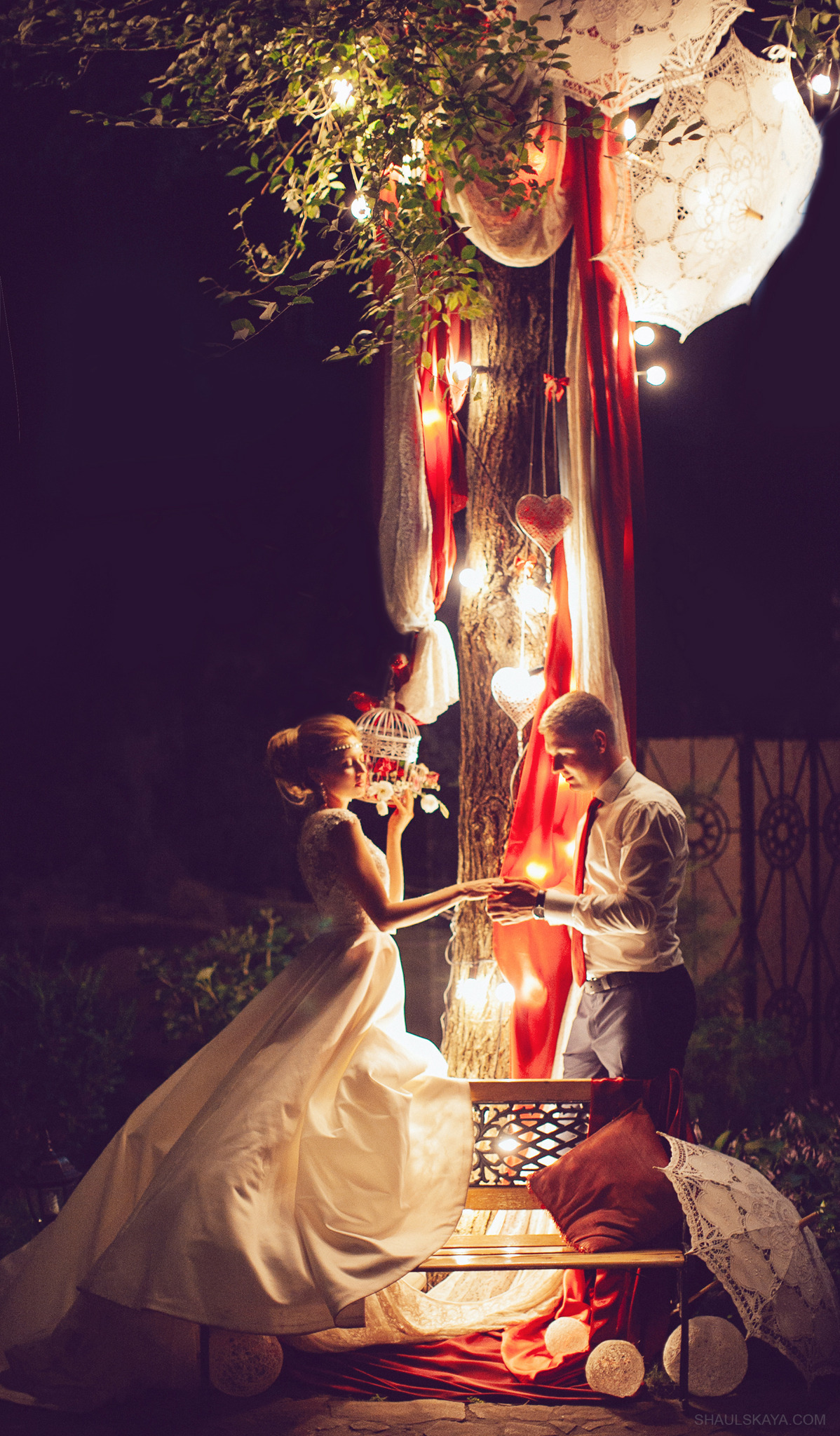 Para narzeczonych w wieczorowej scenerii: panna młoda w białej sukni trzyma klatkę z kwiatami, pan młody w koszuli i krawacie, oboje stoją przy drzewie udekorowanym lampkami, czerwonymi wstążkami...