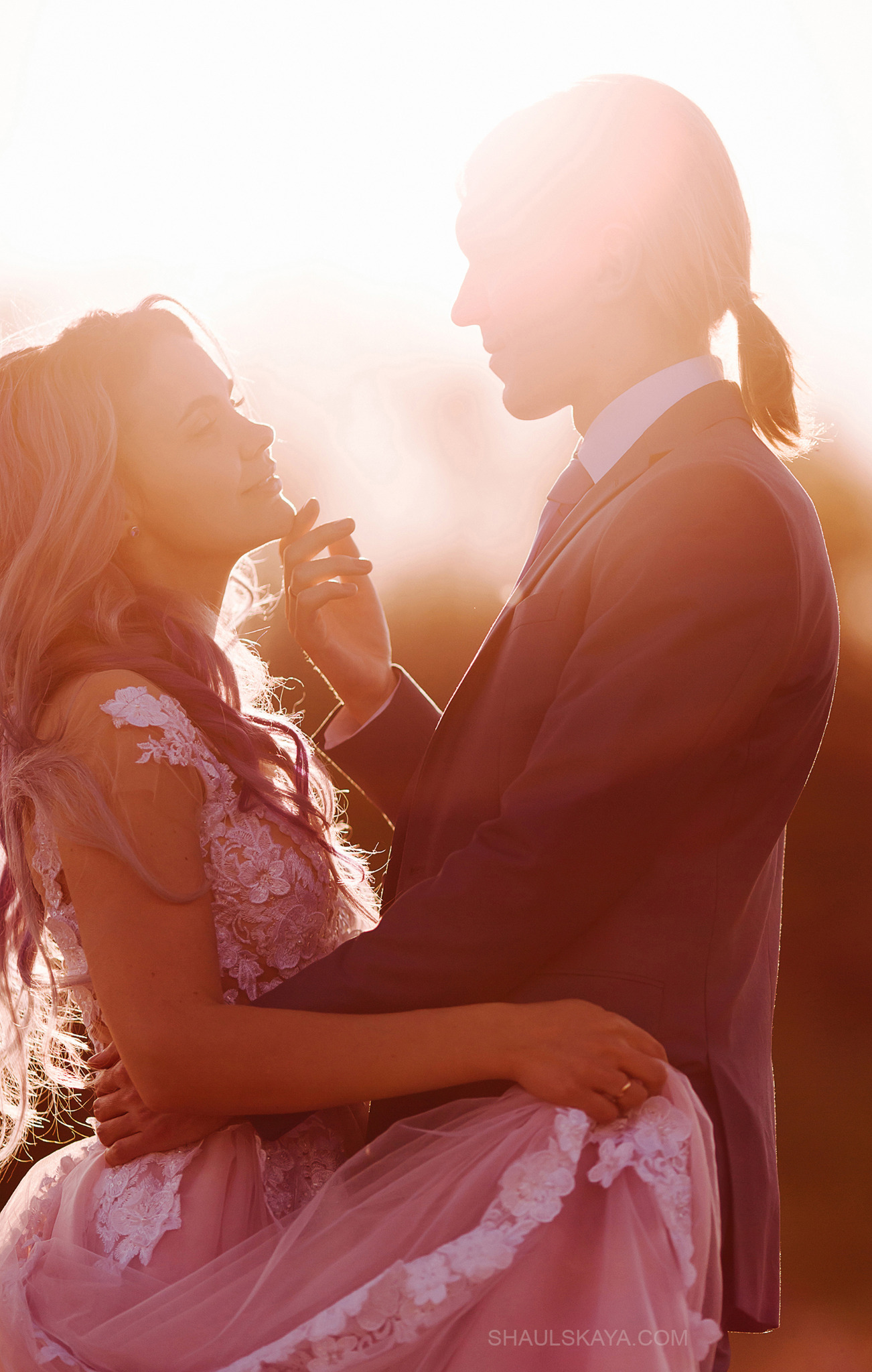 Para w objęciach, kobieta w koronkowej sukni z tiulowym dołem, mężczyzna w garniturze, delikatne promienie zachodzącego słońca tworzą romantyczną aurę, włosy kobiety z fioletowymi pasemkami.