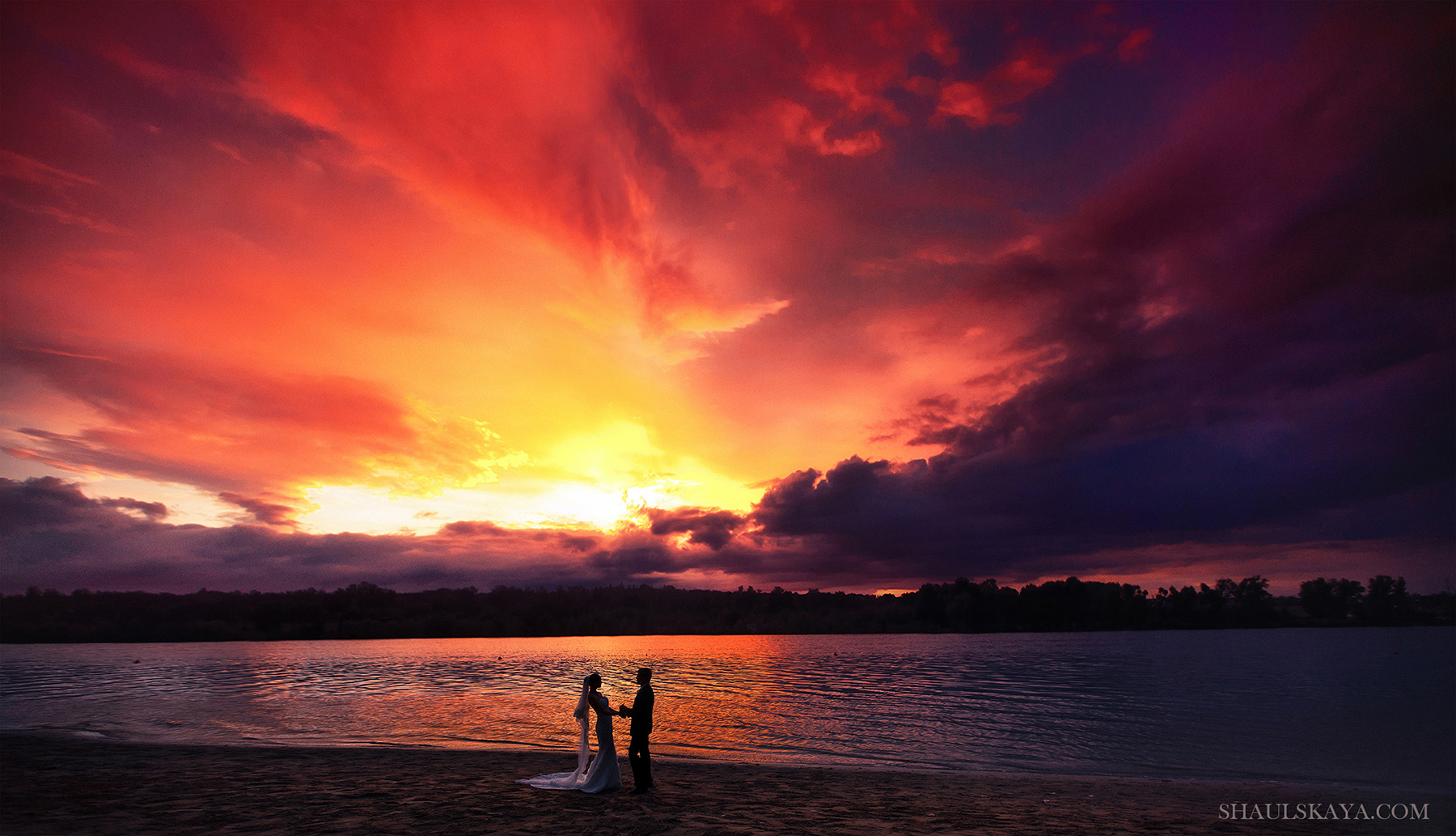 Para trzymająca się za ręce na plaży o zachodzie słońca, dramatyczne niebo w odcieniach czerwieni i fioletu odbija się w wodzie.
