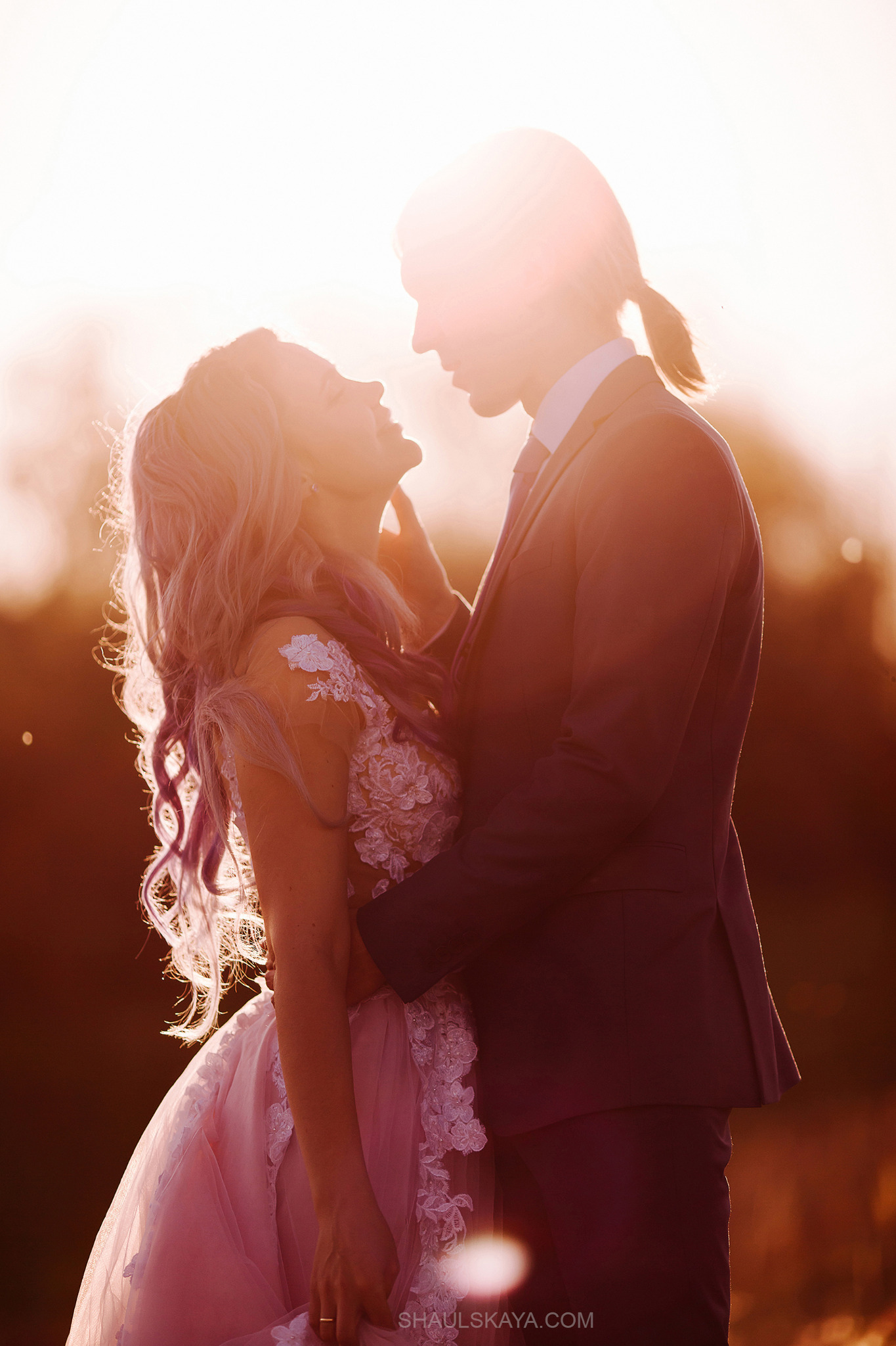 Para zakochanych w kontraście z zachodzącym słońcem: ona w sukni ślubnej z fioletowymi włosami, on w garniturze, delikatnie się obejmują.