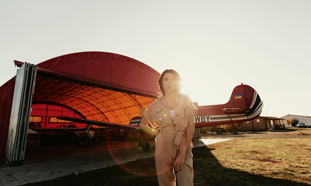 Kobieta w beżowym kombinezonie pozuje na tle czerwonego samolotu z ukraińską flagą na ogonie i otwartego hangaru w słoneczny dzień.