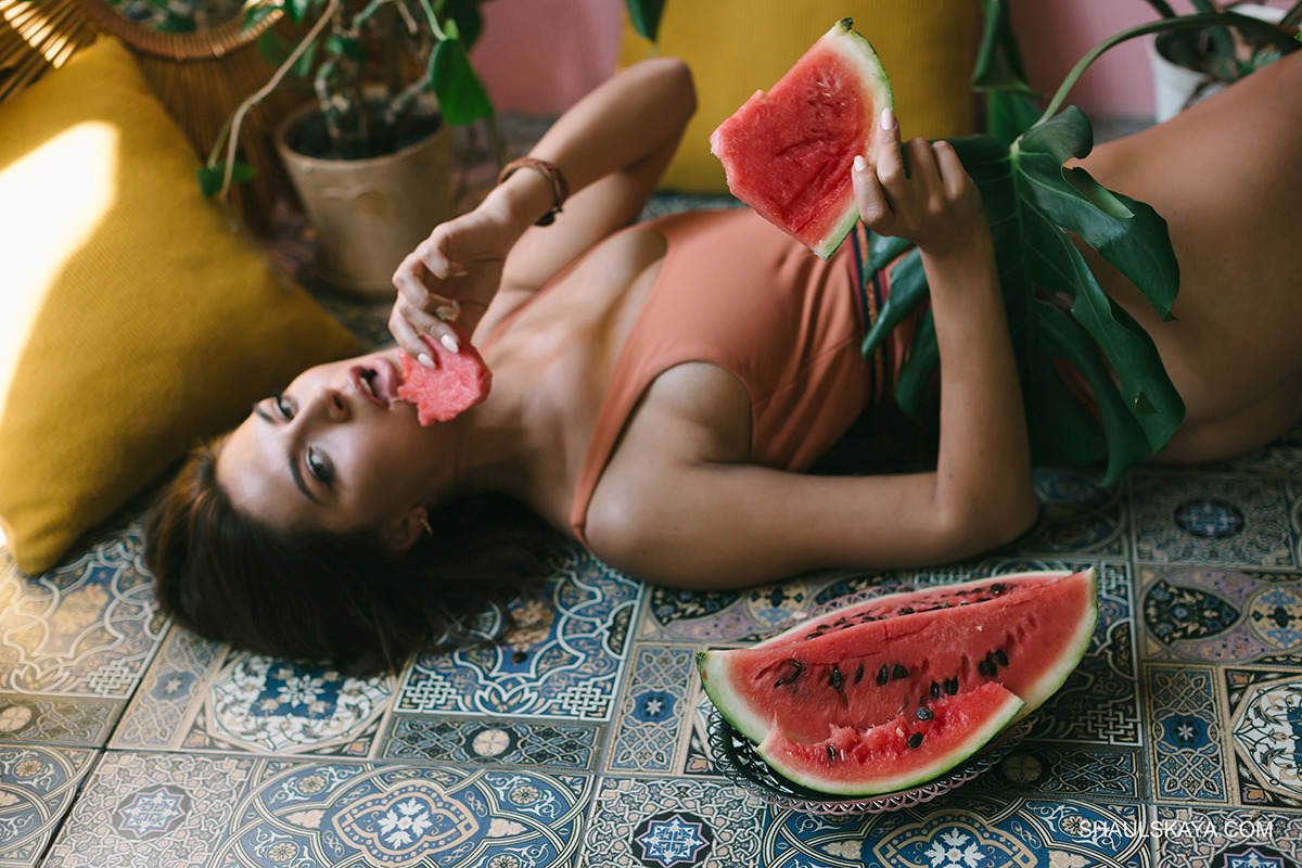 Kobieta leżąca na wzorzystej podłodze w stroju kąpielowym, trzyma kawałek arbuza przy ustach, obok liść monstery i połówka arbuza na talerzyku, żółta poduszka w tle.