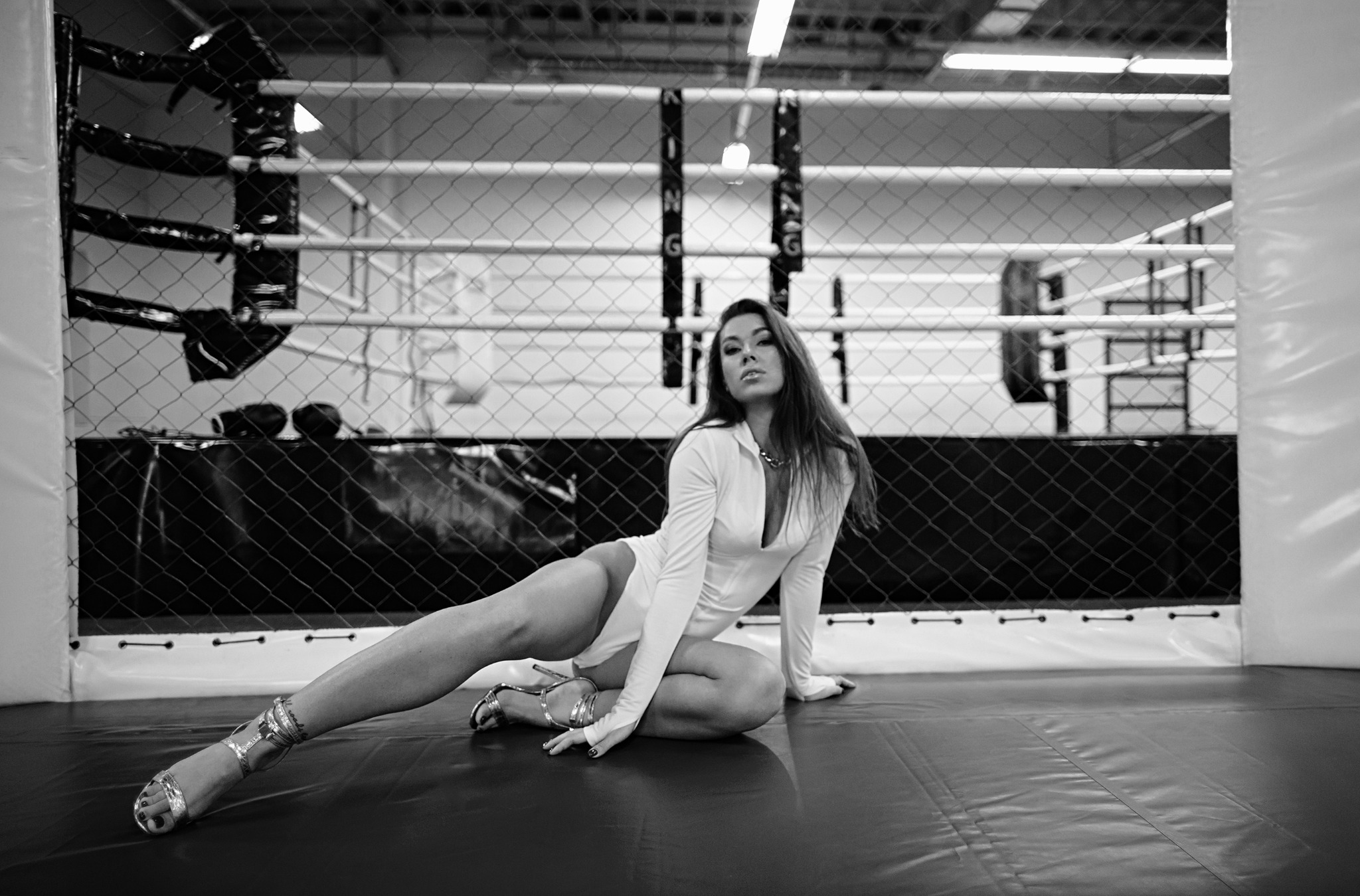 Czarno-białe zdjęcie kobiety w białym body i srebrnych szpilkach, siedzącej na podłodze ringu bokserskiego, z siatką ringu w tle.