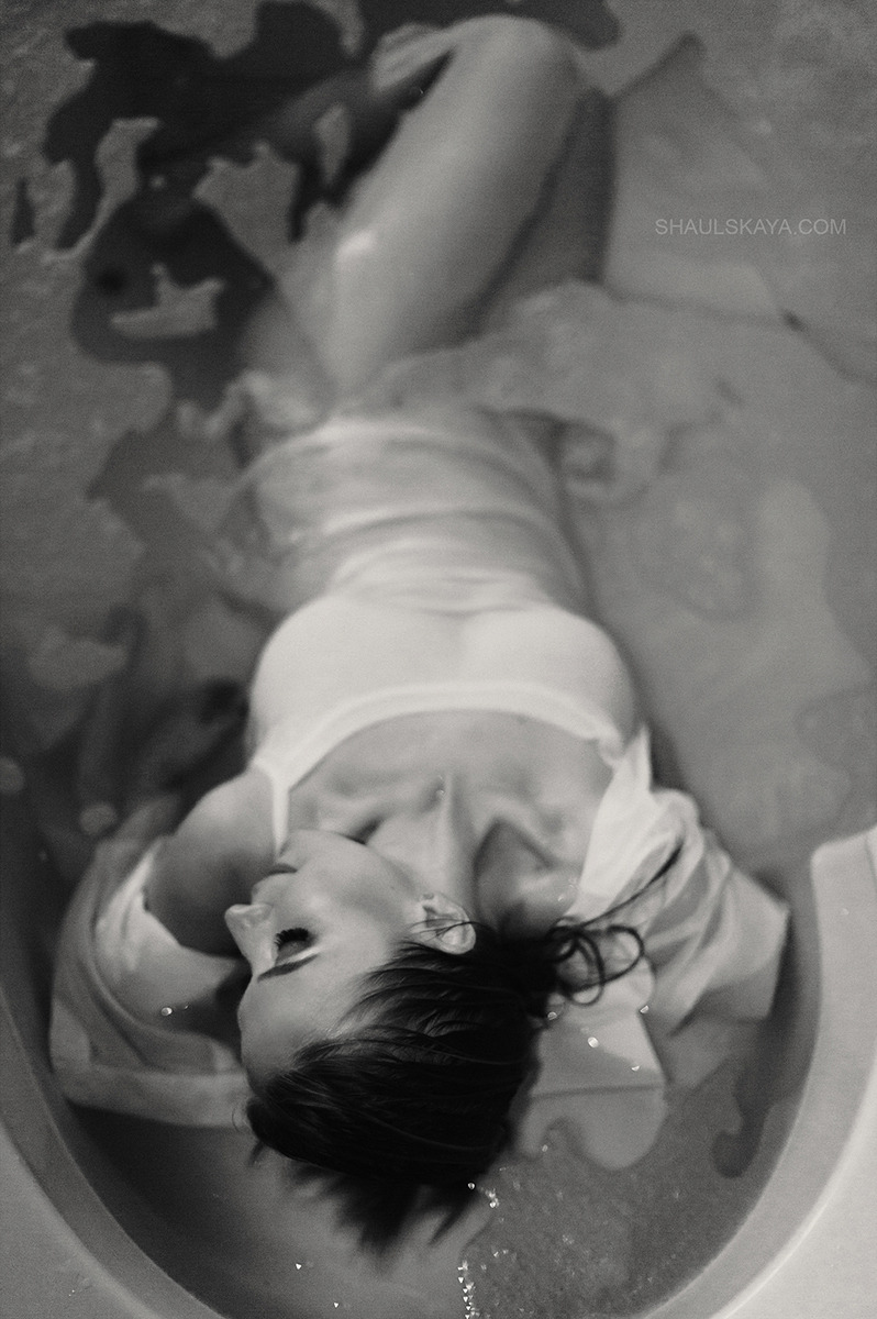 Czarno-białe ujęcie kobiety leżącej w wannie wypełnionej wodą, z zamkniętymi oczami i delikatnym uśmiechem, skierowana głową w lewo, w tle widoczne refleksy światła na wodzie.