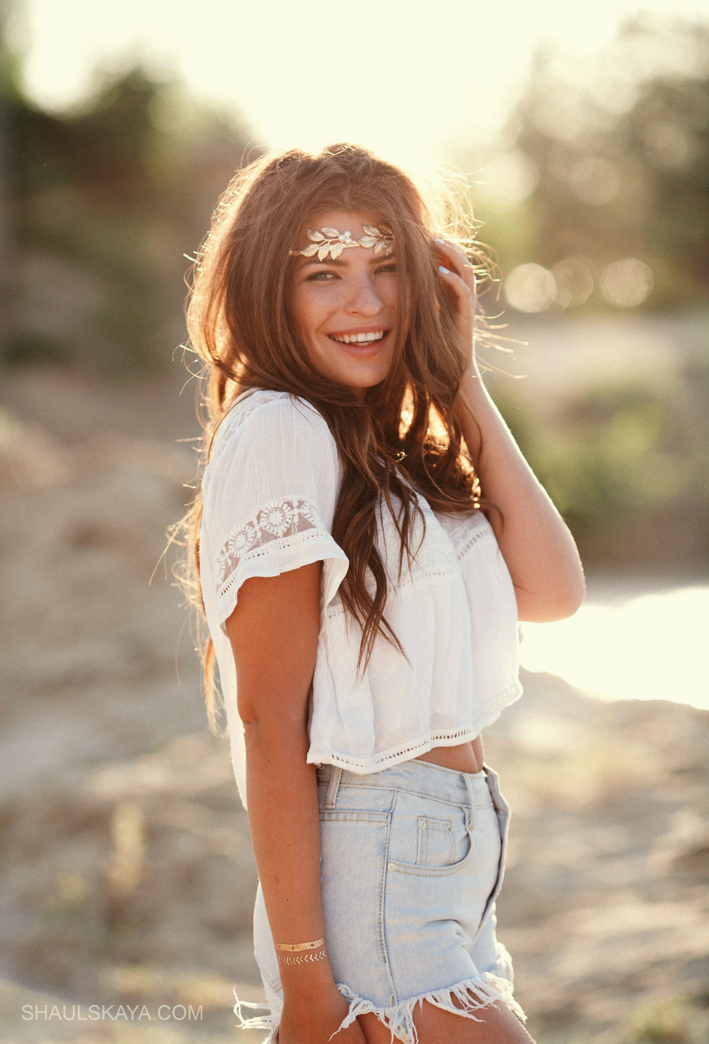 Uśmiechnięta młoda kobieta z brązowymi włosami, w białej bluzce z koronkowymi rękawami i dżinsowych szortach, z ozdobną opaską na głowie, pozująca na tle rozmytego, słonecznego krajobrazu.