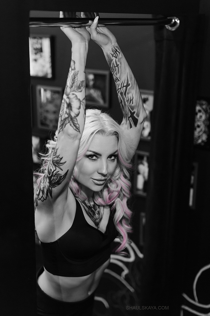 Portret kobiety z tatuażami na ramionach, trzymającej się drążka, z blond włosami z różowymi pasemkami, w czarnym sportowym topie, na czarno-białym zdjęciu z kolorowymi włosami.
