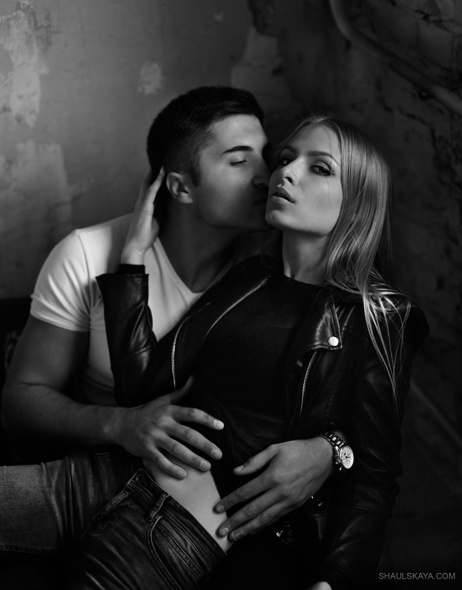 Czarno-białe zdjęcie pary: mężczyzna w białym T-shircie całuje kobietę w skórzanej kurtce, która patrzy w obiektyw z wyrazistym makijażem oczu, na tle ściany z efektem starego tynku.