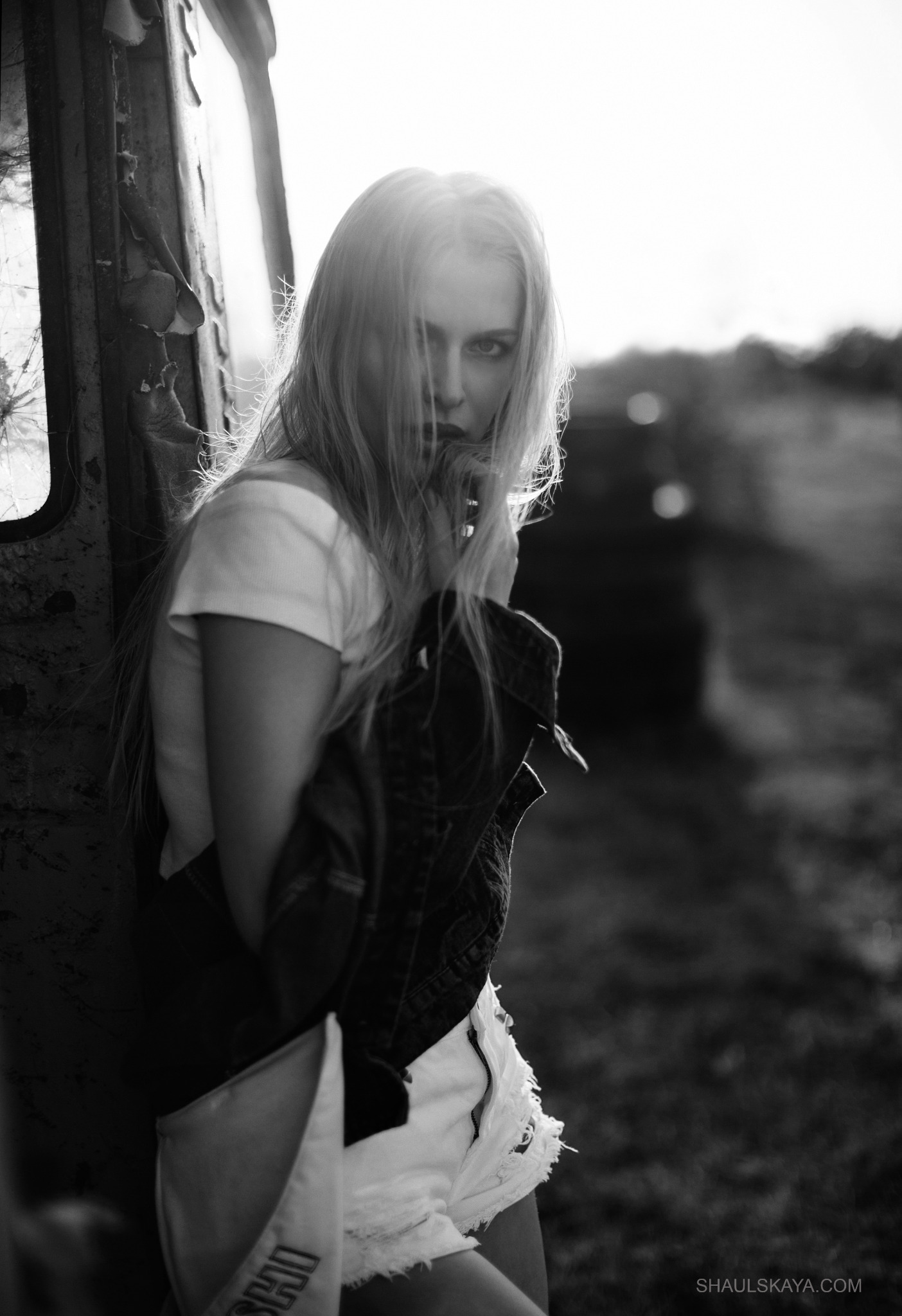 Czarno-białe ujęcie kobiety z blond włosami, opierającej się o zardzewiałą konstrukcję z rozbitym oknem, w tle rozmyty krajobraz, ostre światło słoneczne.