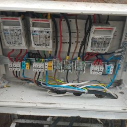 Fest-Instal - Otwarta skrzynka elektryczna z trzema bezpiecznikami APATOR RBK 00, listwami zaciskowymi i przewodami w różnych kolorach, widoczne ślady użytkowania na obudowie.