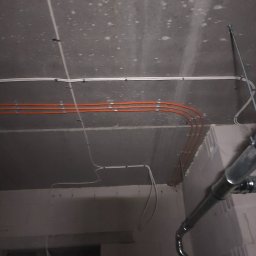 Fest-Instal - Pomarańczowe i białe przewody elektryczne zamocowane klamrami do surowego, betonowego sufitu w trakcie instalacji, widoczna rura wentylacyjna w tle.