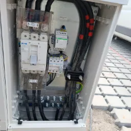 Otwarta skrzynka elektryczna z widocznymi zabezpieczeniami, przewodami i elementami montażowymi; widoczne napisy ostrzegawcze i oznaczenia producentów na elementach. Na górze skrzynki leży klucz...