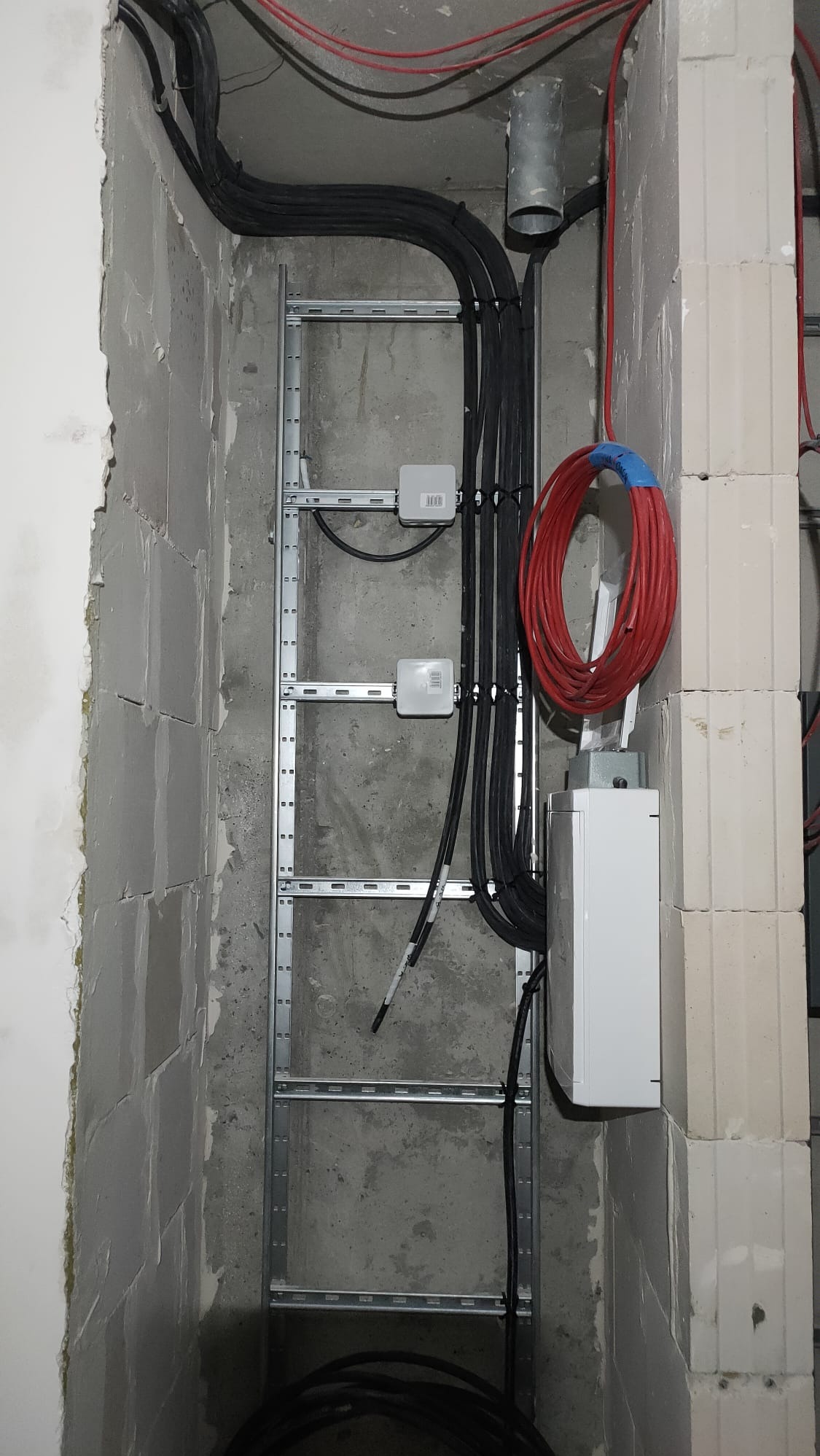 Wewnętrzna instalacja elektryczna w surowym stanie: kable czarne i czerwone prowadzone wzdłuż metalowej drabinki montażowej na ścianie z betonu komórkowego, widoczne puszki instalacyjne i zwój...
