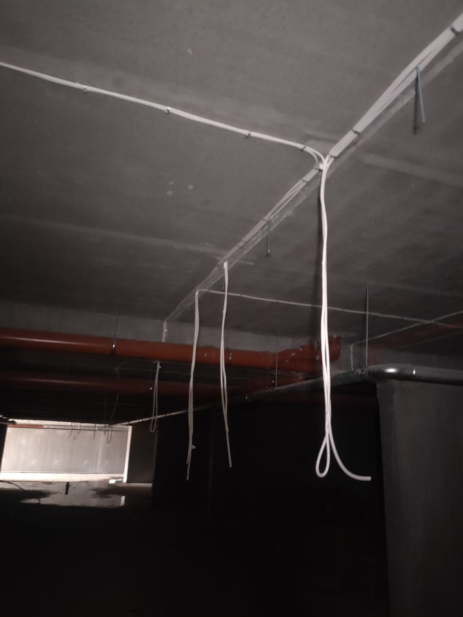 Instalacja przewodów elektrycznych w korytkach pod sufitem, z wiszącymi luźnymi kablami i widocznymi rurami instalacyjnymi w tle, w surowym, ciemnym pomieszczeniu.