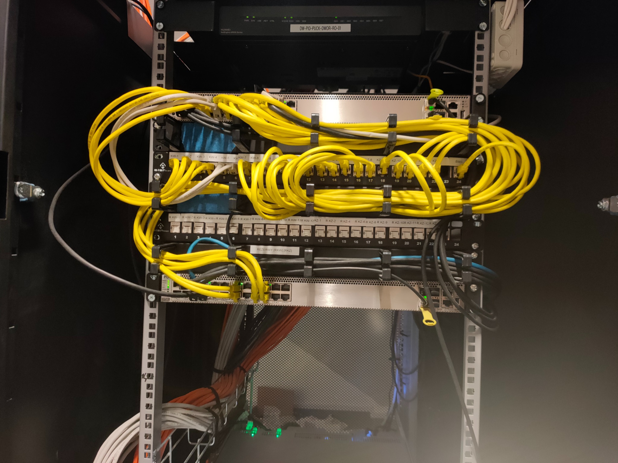Szafa rack z uporządkowanymi kablami sieciowymi, głównie w kolorze żółtym, zabezpieczonymi czarnymi opaskami, widoczne przełączniki sieciowe i patch panele, z etykietami i diodami LED.