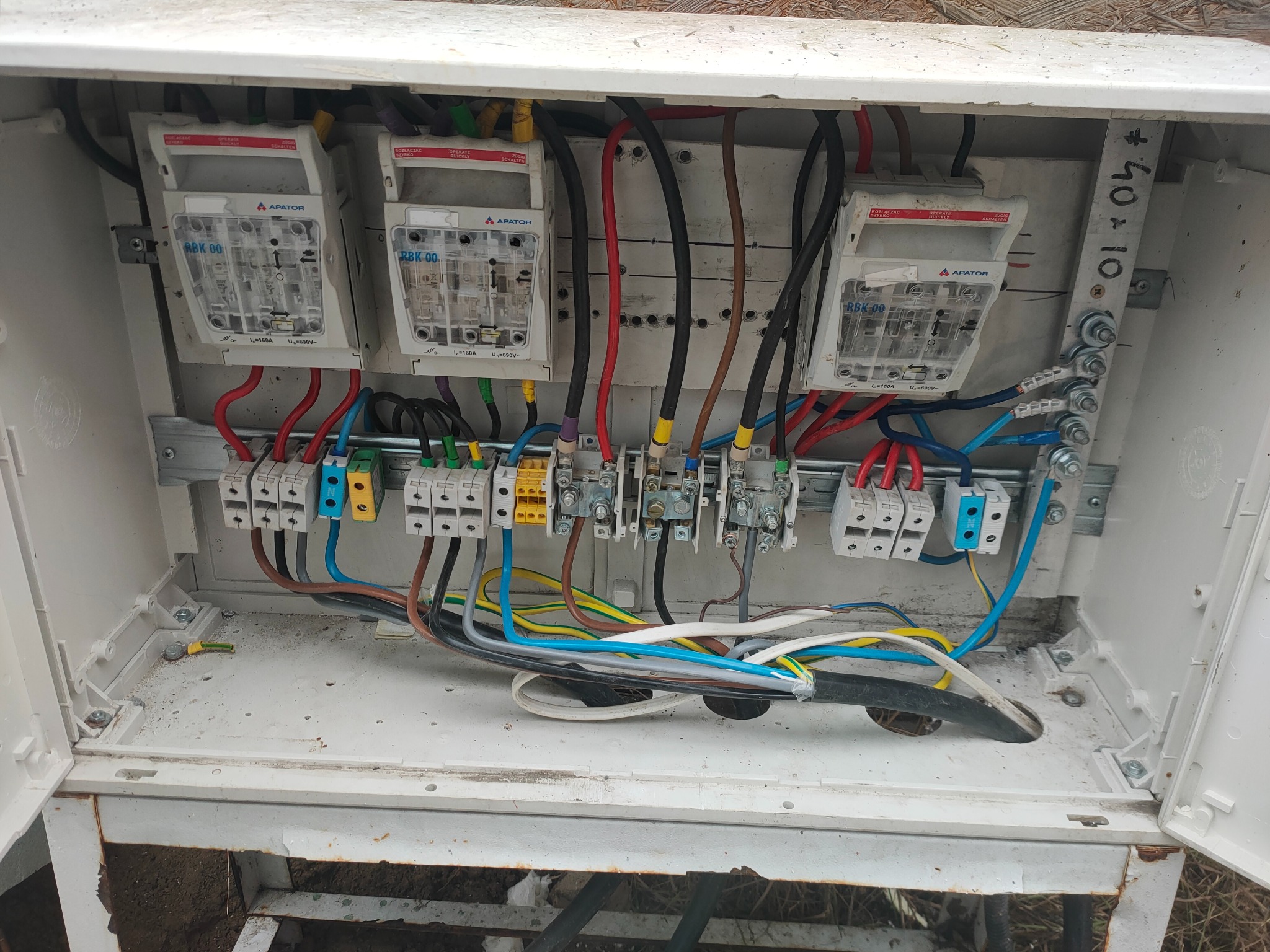 Otwarta skrzynka elektryczna z trzema bezpiecznikami APATOR RBK 00, listwami zaciskowymi i przewodami w różnych kolorach, widoczne ślady użytkowania na obudowie.