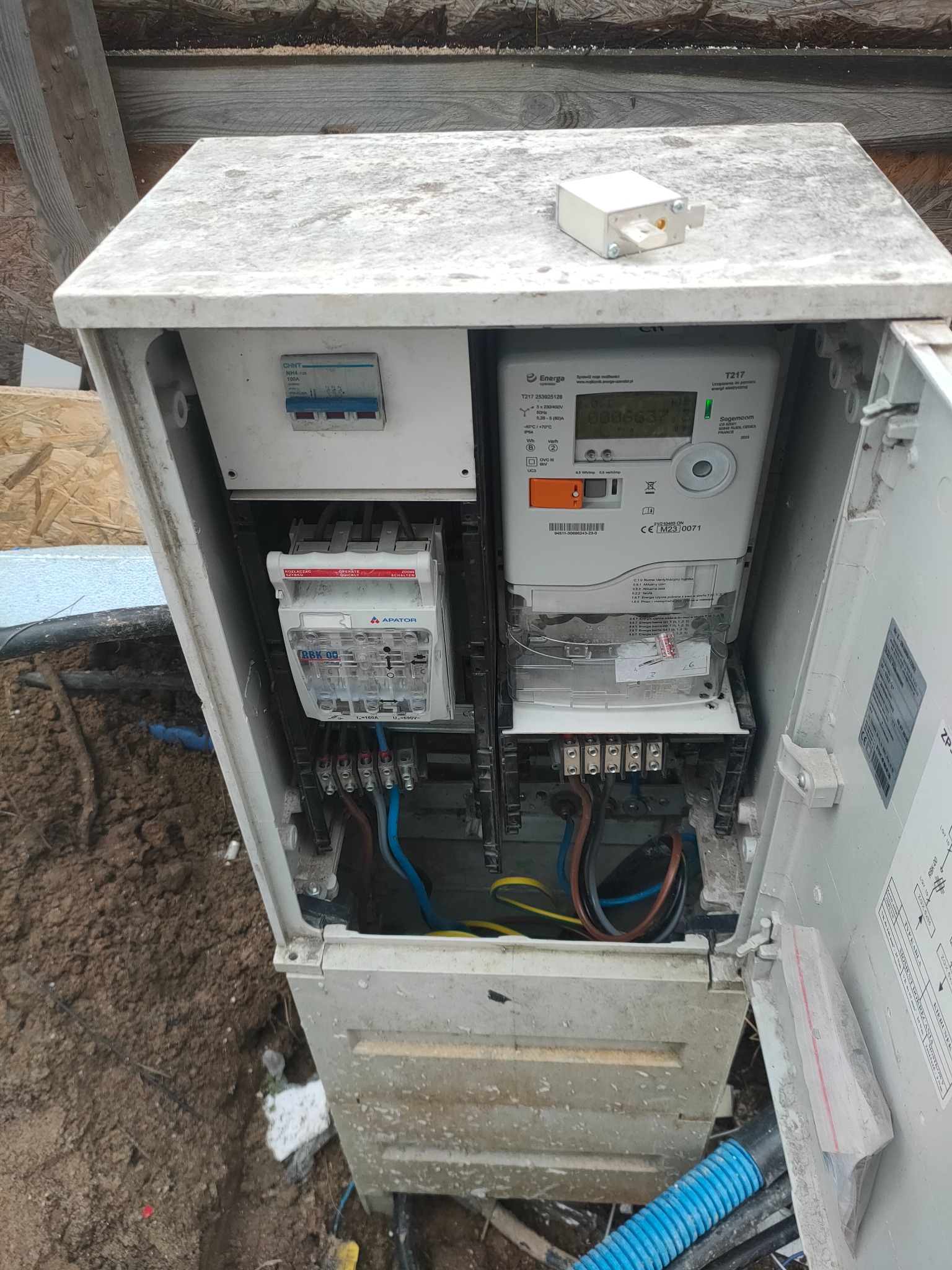 Otwarta skrzynka z licznikiem energii elektrycznej i zabezpieczeniami, widoczne kable, złącza i aparatura pomiarowa, całość lekko zabrudzona ziemią.