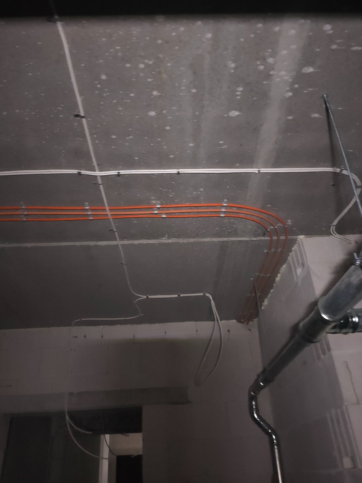 Pomarańczowe i białe przewody elektryczne zamocowane klamrami do surowego, betonowego sufitu w trakcie instalacji, widoczna rura wentylacyjna w tle.