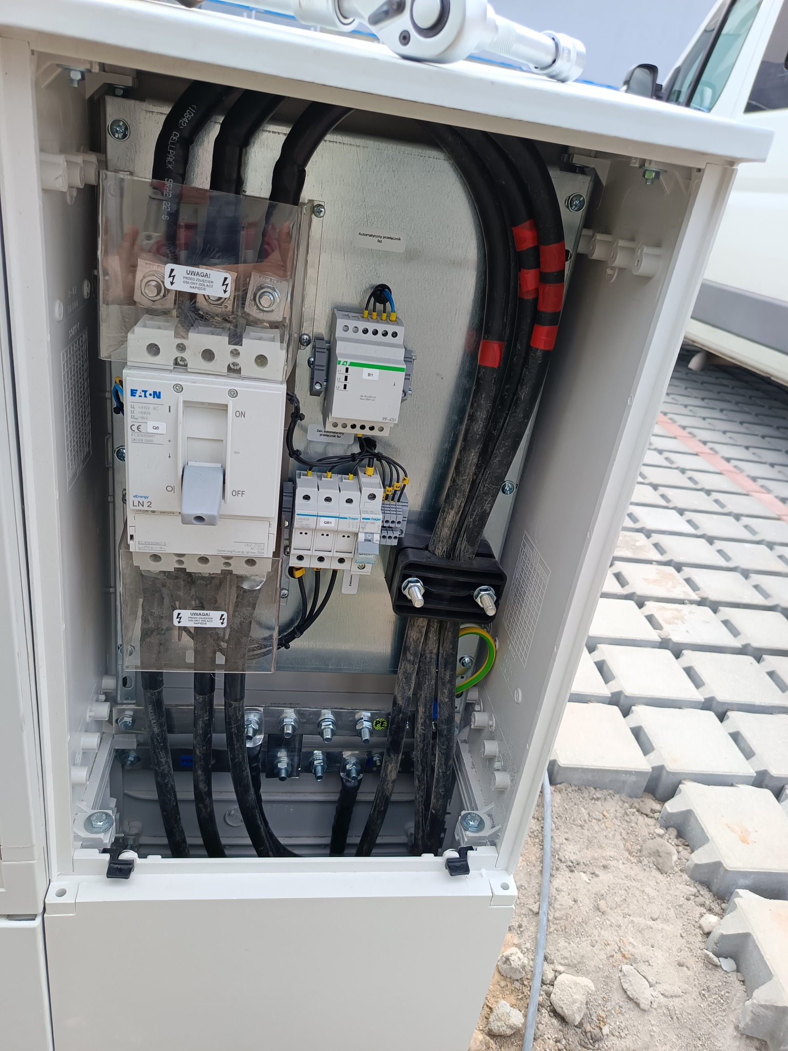 Otwarta skrzynka elektryczna z widocznymi zabezpieczeniami, przewodami i elementami montażowymi; widoczne napisy ostrzegawcze i oznaczenia producentów na elementach. Na górze skrzynki leży klucz...