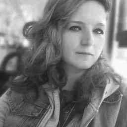 Portret kobiety w dżinsowej kurtce z kapturem, czarno-białe zdjęcie z rozmytym tłem, widoczny wisiorek z motywem drzewa.