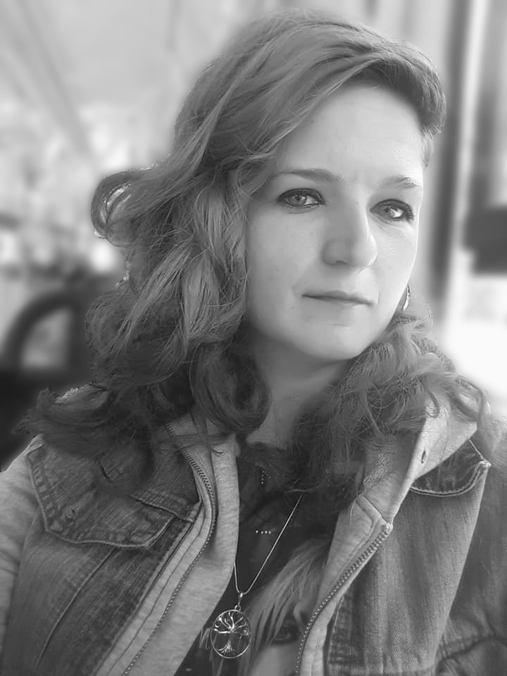 Portret kobiety w dżinsowej kurtce z kapturem, czarno-białe zdjęcie z rozmytym tłem, widoczny wisiorek z motywem drzewa.